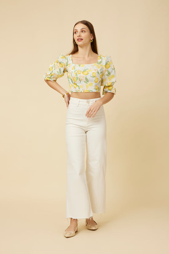 Model stands confidently with hands on hips in the Citrus Dream Crop Top, featuring a bright lemon print, flattering princess cut, and puff sleeves, paired with white high-waisted pants for a refreshing daytime ensemble.