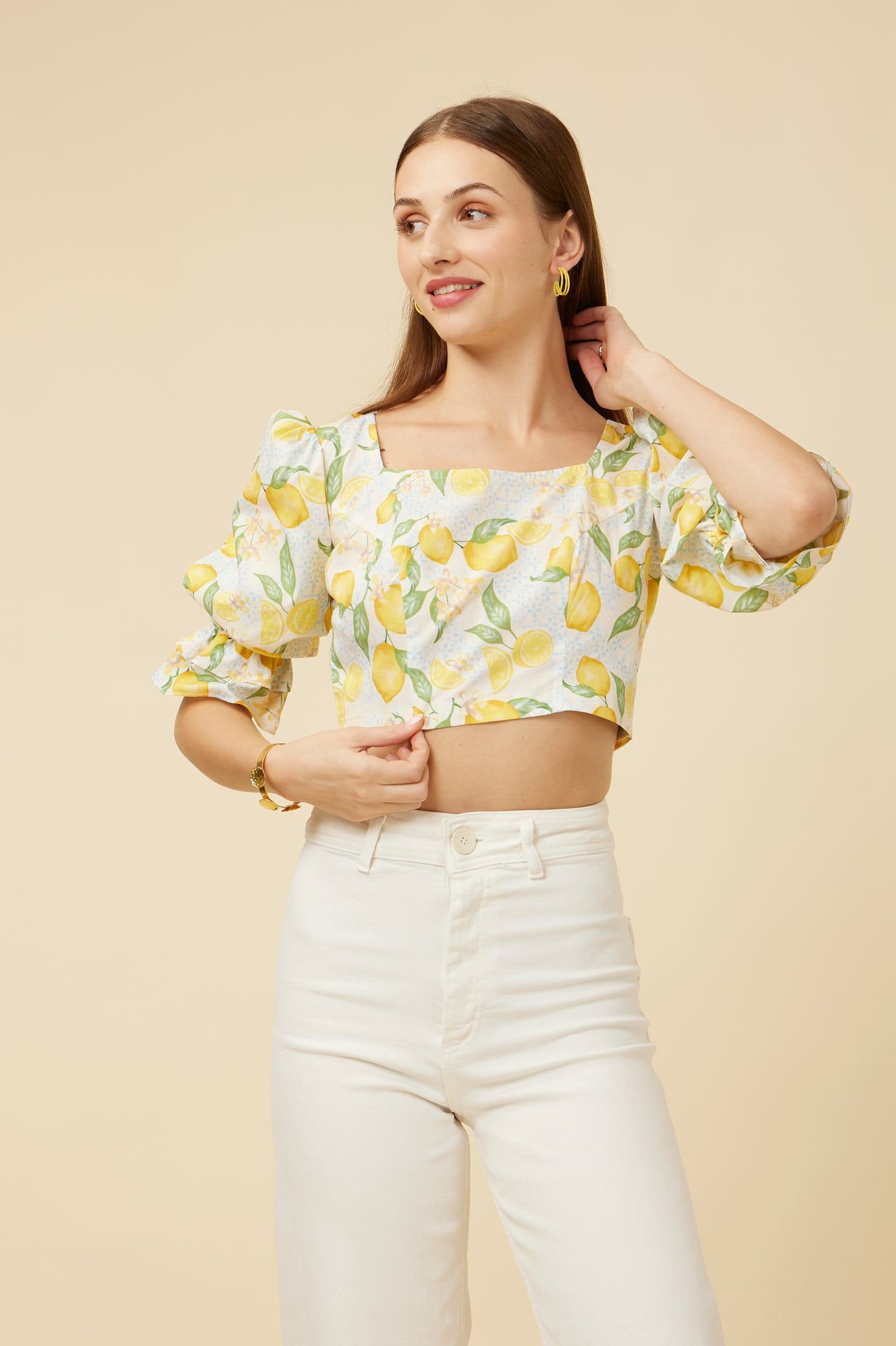 Model touches her hair, showcasing the Citrus Dream Crop Top's rectangle neckline and vibrant lemon print, complemented by puff elbow-length sleeves with playful frill ends, exuding a fresh and summery vibe.