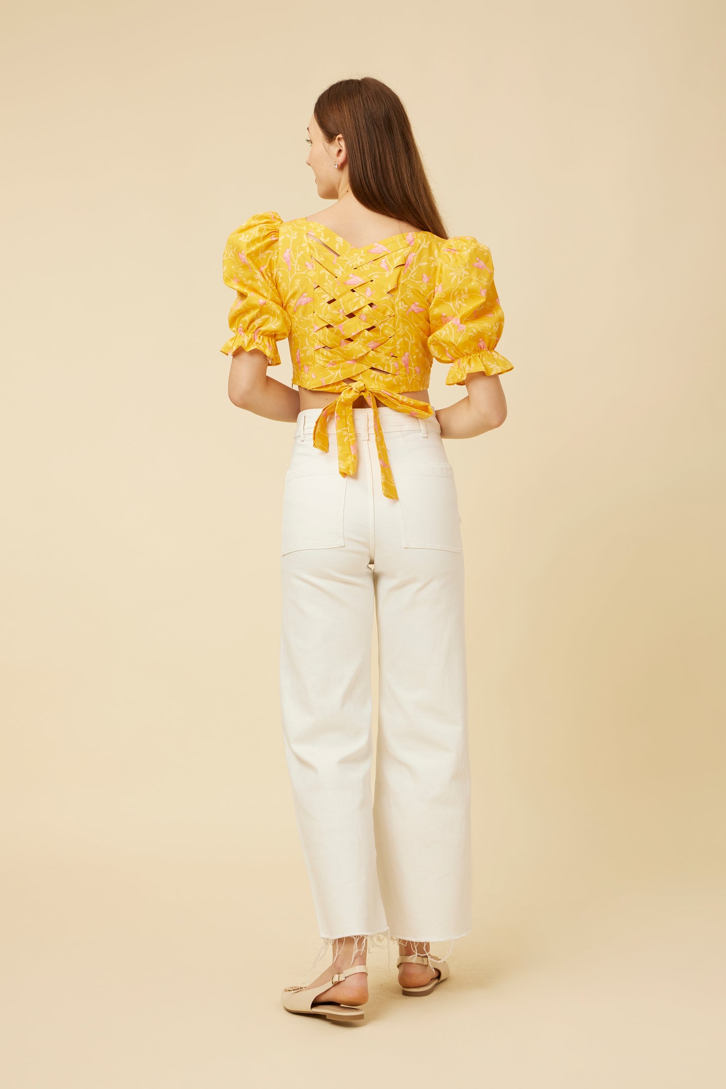 Rear view of the Peela Sunshine Crop Top displaying the criss-cross and tie-back details, paired with high-waisted white pants on a model