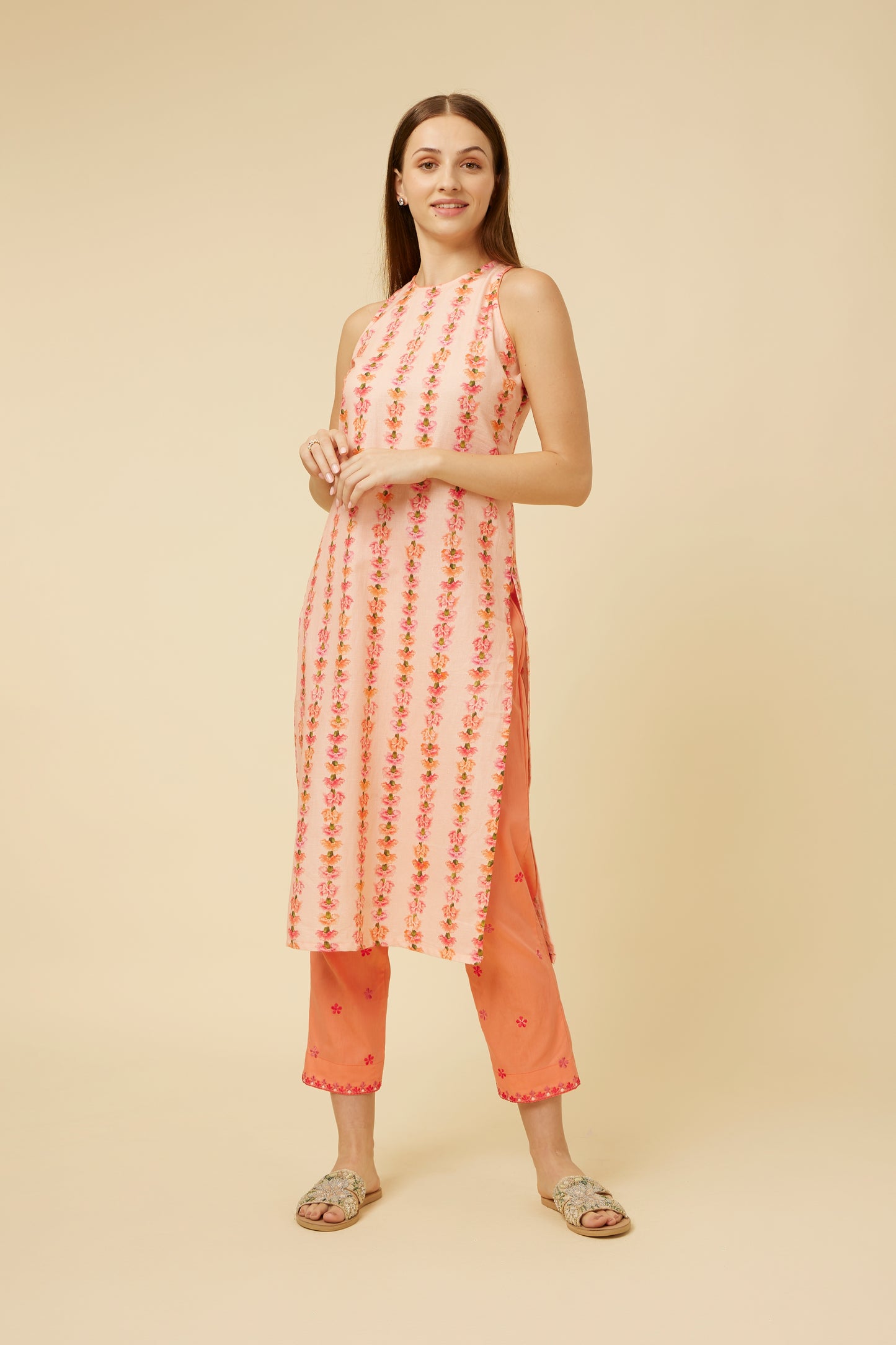 A model poses in a sleeveless, semi-halter neck kurta in a soft pink hue with vertical stripes of delicate carnation embroidery. The kurta's A-line silhouette is complemented by a side slit, revealing matching pink pants with subtle embroidery at the hem