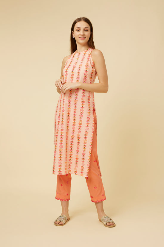 A model poses in a sleeveless, semi-halter neck kurta in a soft pink hue with vertical stripes of delicate carnation embroidery. The kurta's A-line silhouette is complemented by a side slit, revealing matching pink pants with subtle embroidery at the hem