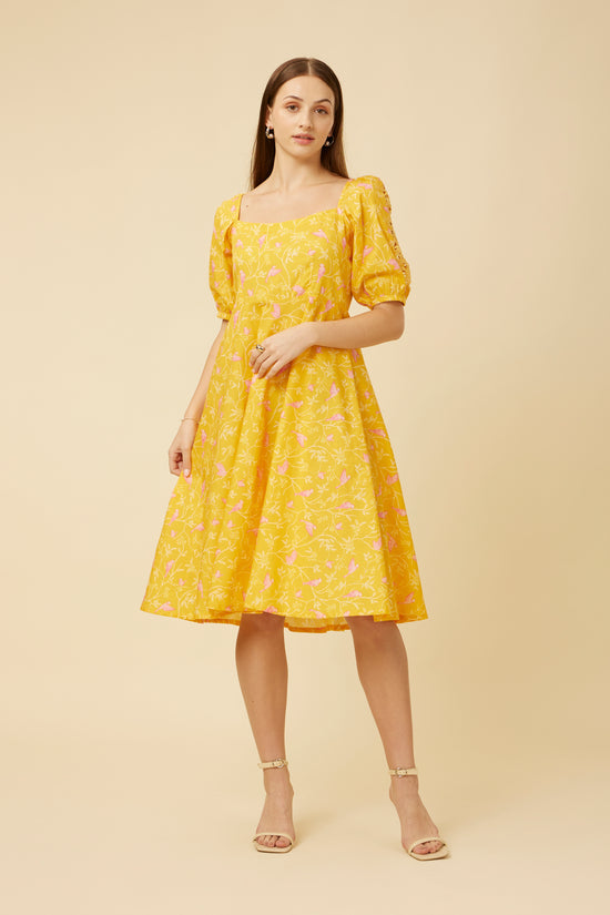 Model in Peela Sunshine Dress with a wide rectangle neckline and cutwork detailing on the sleeves, radiating joy in a cheerful yellow fabric adorned with pink birds