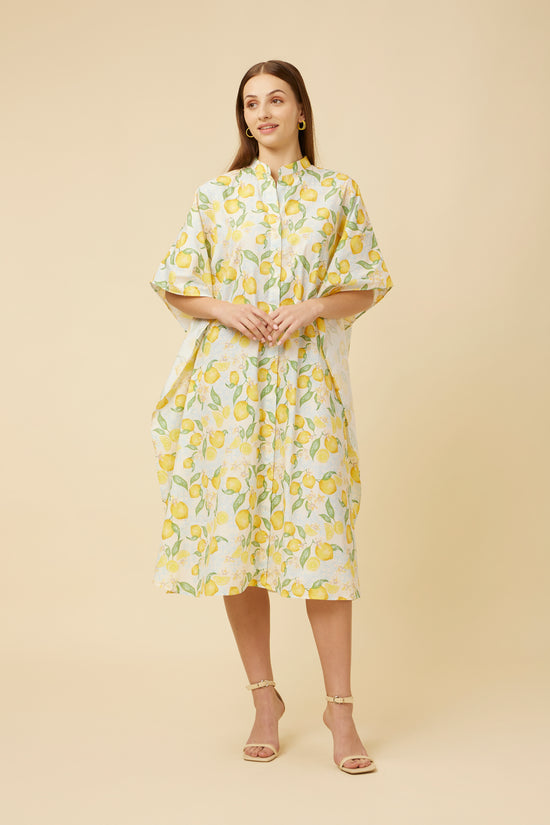Model clasps her hands in front of the Citrus Dream Shirt Dress, showcasing the collar neckline and the lively lemon print that flows throughout the kaftan, embodying a breezy, summery elegance.