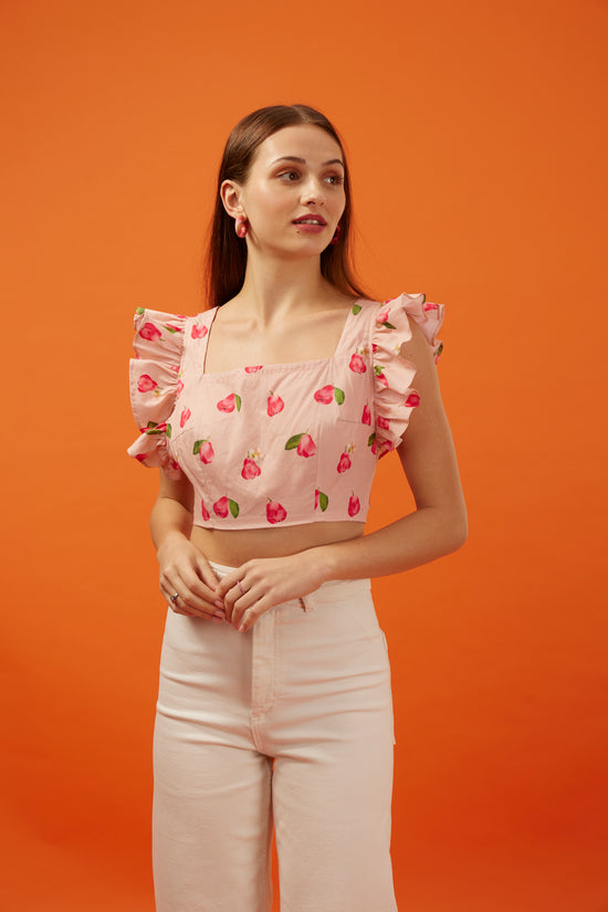 Elegant young woman posing in a Hypsway pastel pink crop top adorned with vibrant tulip prints and ruffled sleeves, paired with sleek light pink trousers, against a warm orange background, capturing the playful yet sophisticated style of Hypsway's crop top collection for the trendy urban woman.