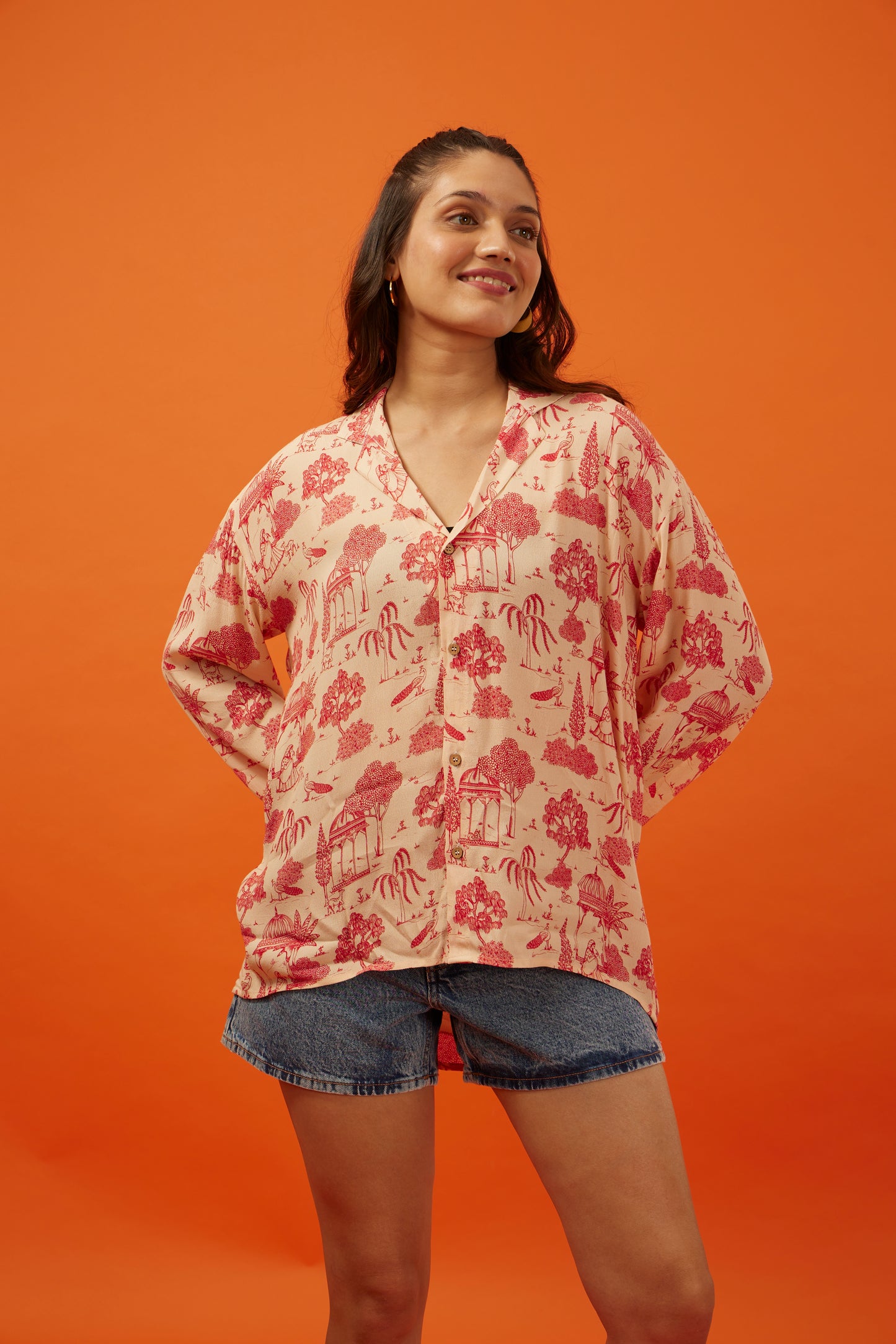 Chic young woman sporting a Hypsway oversized bohemian blouse with intricate Indian palace print in shades of coral and beige, paired with casual denim shorts, exuding a relaxed yet fashionable vibe perfect for a versatile wardrobe.