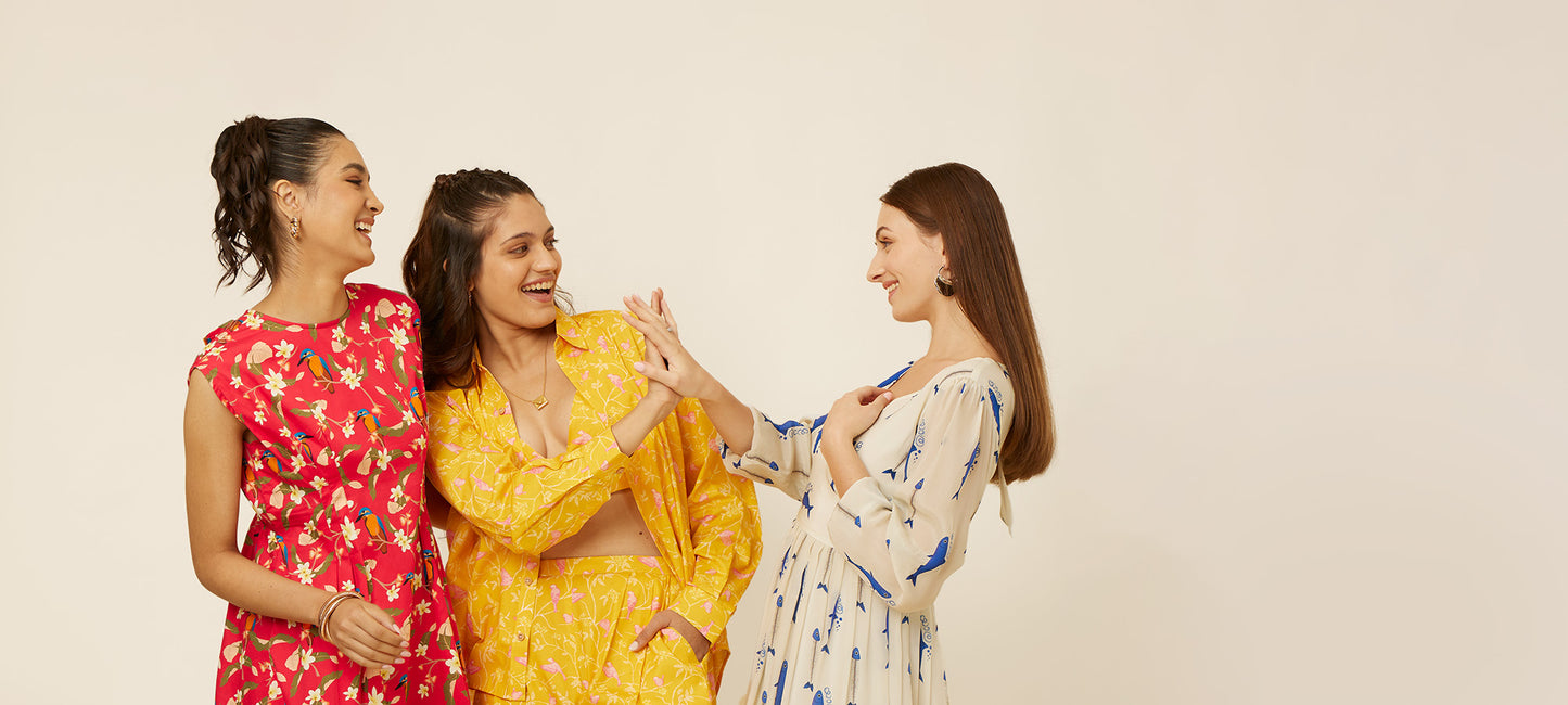 Three fashionable young women in their twenties wearing Hypsway's latest custom-designed apparel; featuring a bold red floral dress, a sunny yellow floral co-ord set, and an elegant white dress with playful blue bird patterns, perfect for the modern Indian woman's chic and versatile wardrobe.